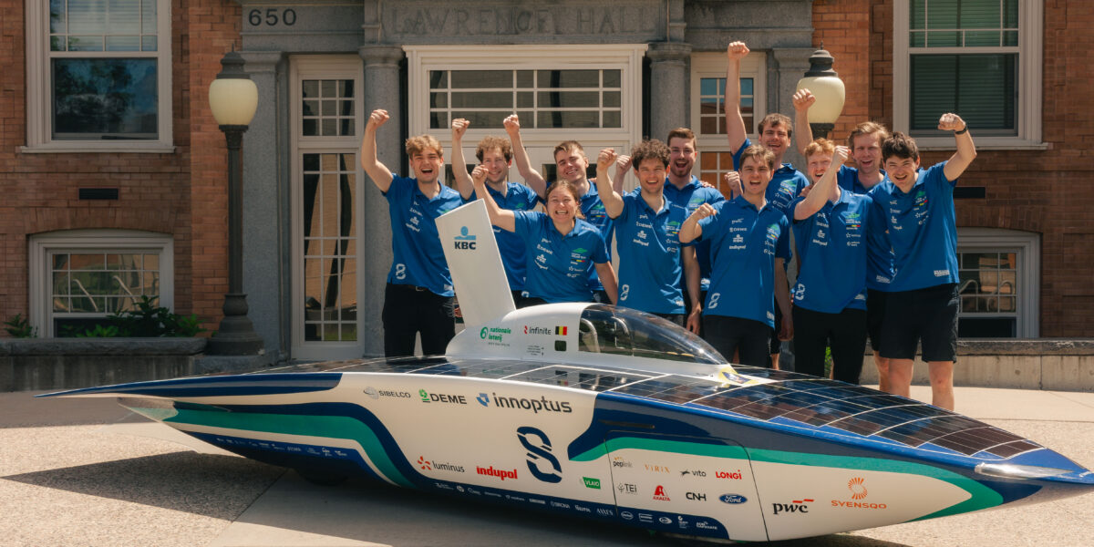 A group of 11 people in blue polos lift their arms for a photo while standing next to a solar-powered car
