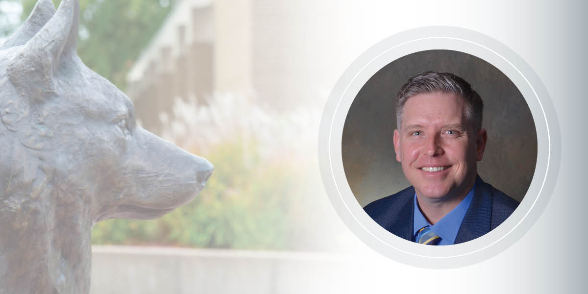 A man in a blue suit and tie smiles for a headshot next to the Husky Plaza statue