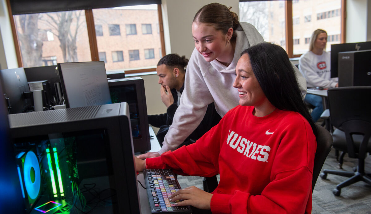 Two women look at a computer screen while students work in the background