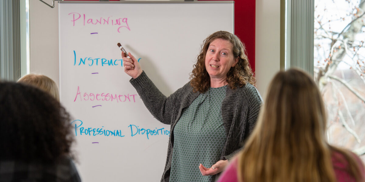 A woman in a grey sweater points to a whiteboard with a class of students out of focus