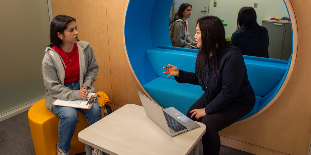 One woman sits on a yellow stool while holding a clipboard, listening to another woman talk with a laptop in front of her