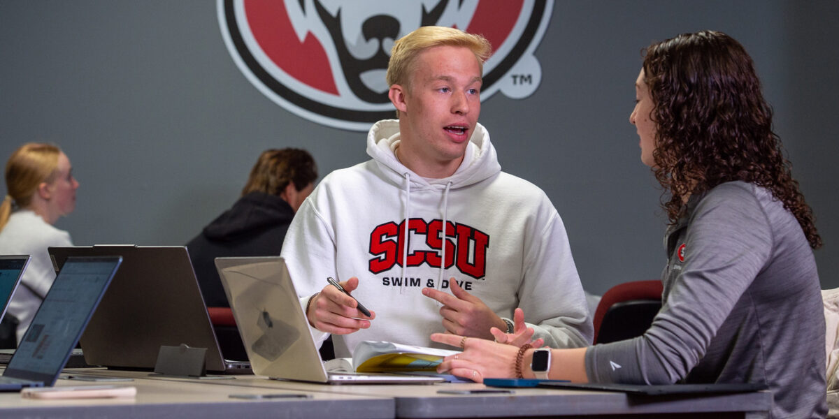 A man in a white SCSU Swim & Dive sweatshirt talks to a woman in a grey quarter zip at a table with laptops visible