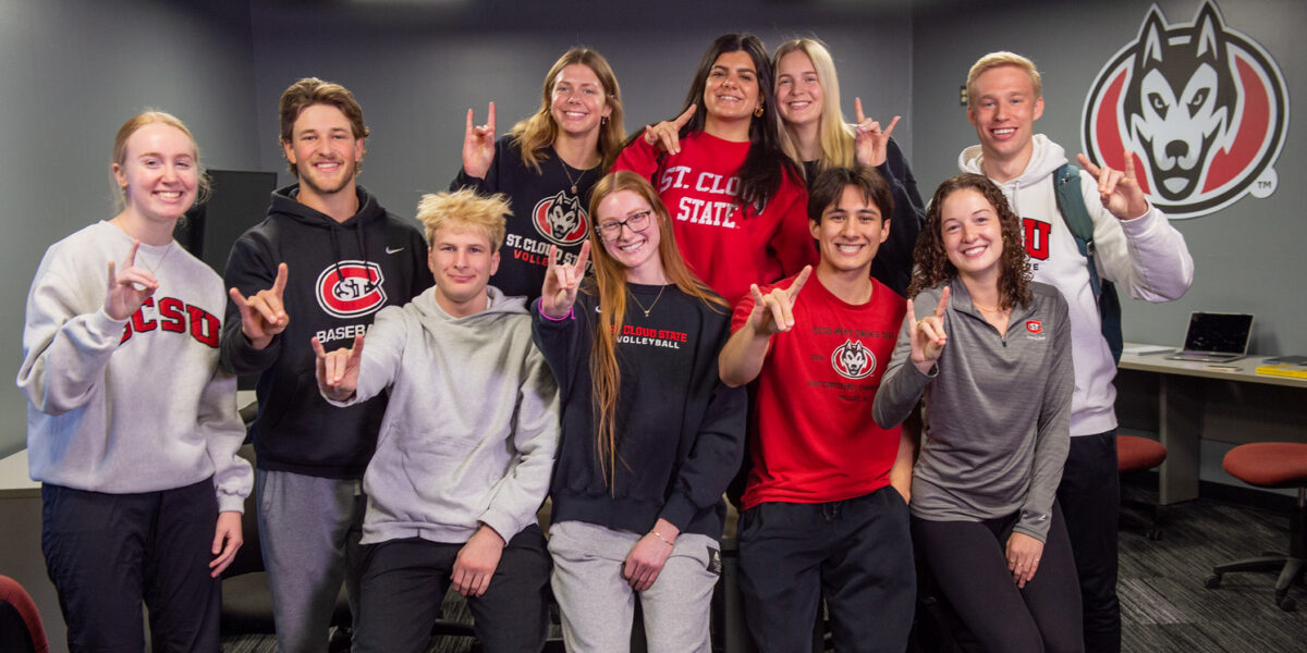 A group of 10 students is SCSU shirts smile at the camera while making the sign of the Husky with their hands