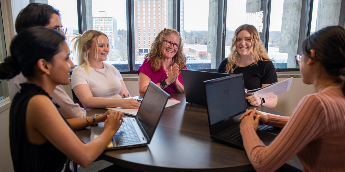 A group of six individuals smile during a round table discussion while working on laptops