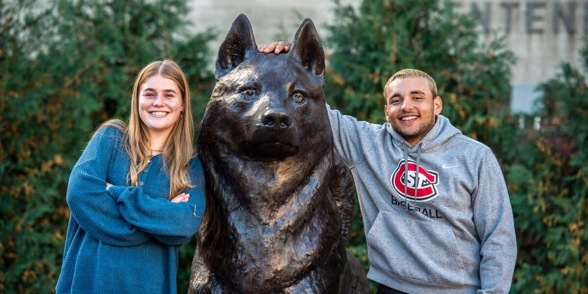 A man and woman smile at the camera while standing next to the Blizzard statue in Husky Plaza