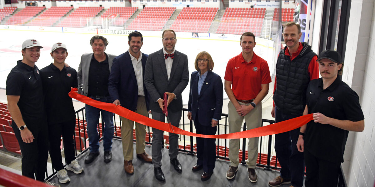 9 people gather together for a ribbon cutting in front of the main ice sheet in the Herb Brooks National Hockey Center